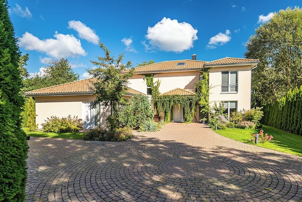 A two-story house with a paved driveway leading to the entrance, surrounded by greenery and a clear blue sky.