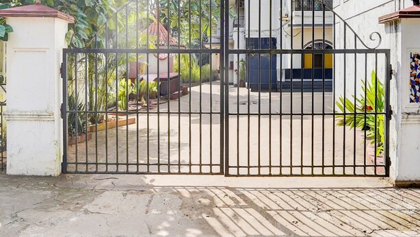 A gated entrance with a metal gate, a paved driveway, and a building with a white facade and blue accents. - Super Townhouse Calangute (Calangute)