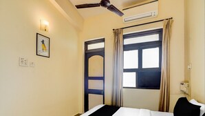A hotel room with a bed, a ceiling fan, an air conditioning unit, a window with curtains, and a wall-mounted light fixture. - Super Townhouse Calangute (Calangute)
