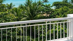 Un balcon doté d’une balustrade blanche, donnant sur une plage tropicale aux eaux d’un bleu limpide et bordée de palmiers.