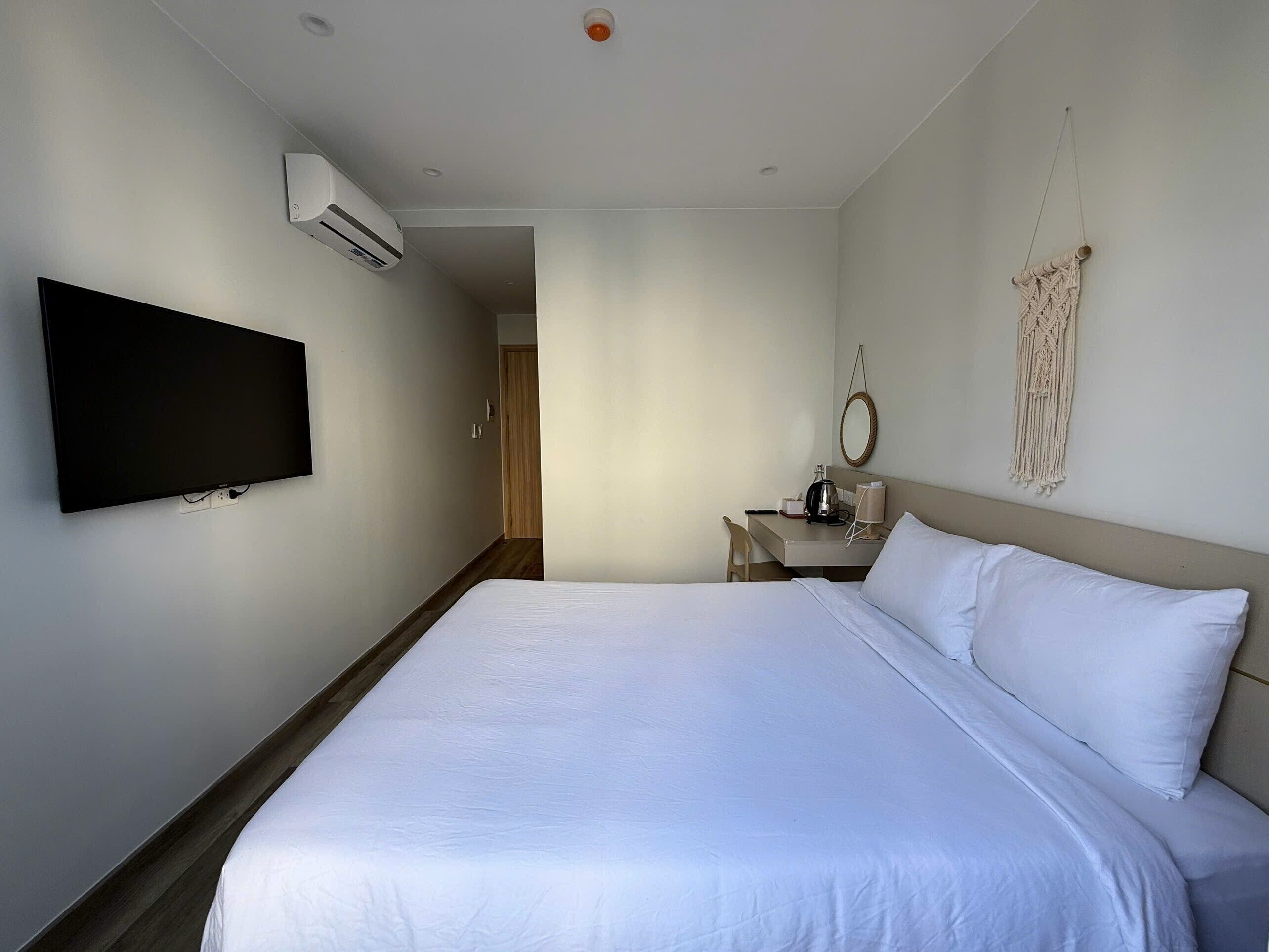 A hotel room with a bed, a flat-screen TV mounted on the wall, a desk with a chair, a macrame wall hanging, and a ceiling-mounted air conditioning unit.