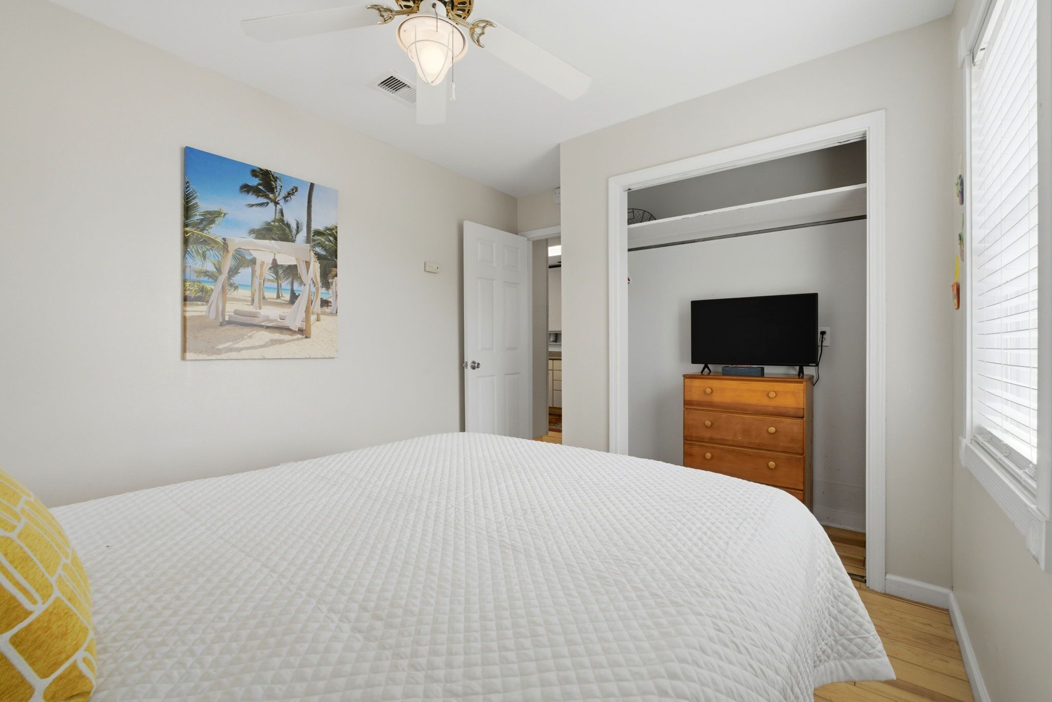 A bedroom with a bed, a ceiling fan, a TV, a wardrobe, and a painting of a beach scene.