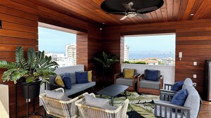 Ein modernes Interieur mit Holzbalkendecke, Deckenventilator, einer Sitzecke mit Sofas und Sesseln, einem Couchtisch und großen Fenstern mit Blick auf die Stadt.