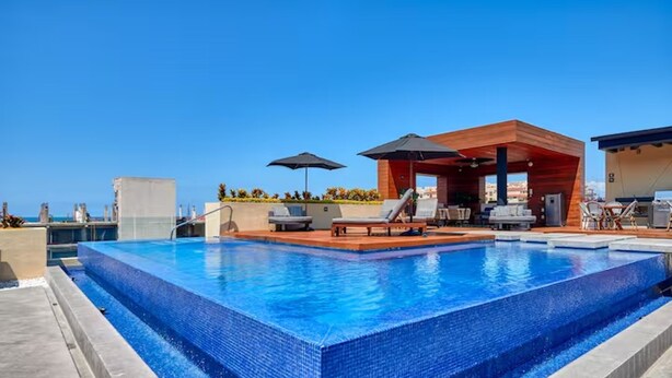 Ein rechteckiger Swimmingpool mit einer gefliesten Kante, umgeben von Liegestühlen und Sonnenschirmen, sowie einer Holzterrasse.