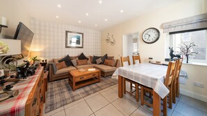 A living room with a sofa, coffee table, dining table, and chairs. There is a clock on the wall and a window with a view. - Fionas Cottage -  a house that sleeps 6 guests  in 3 bedrooms (Crieff)