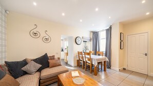 A living room with a sofa, a wooden coffee table, a dining area with a table and chairs, a clock on the wall, and a door. - Fionas Cottage -  a house that sleeps 6 guests  in 3 bedrooms (Crieff)