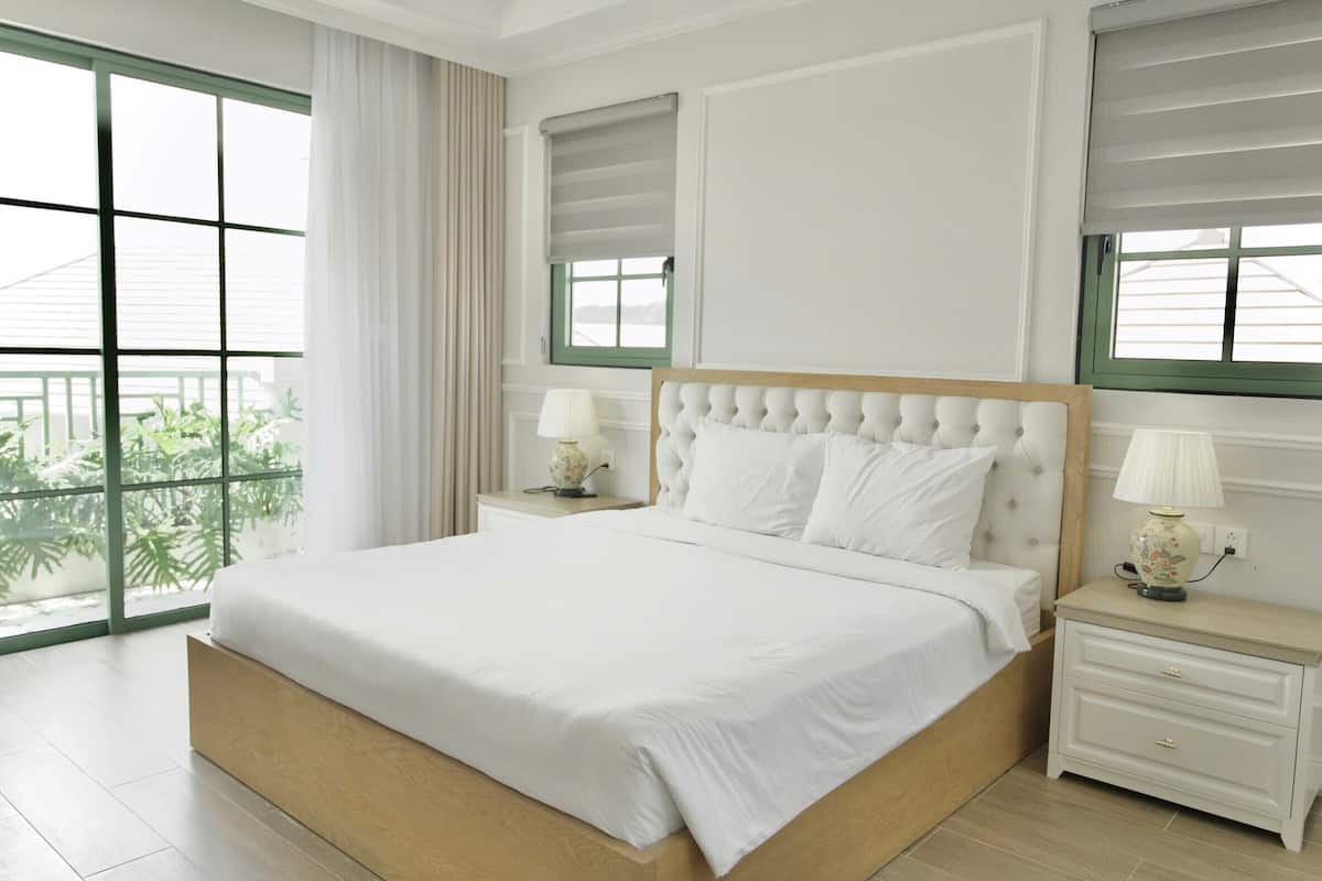 Una cama grande con cabecera de madera, dos mesitas de noche con lámparas, y vistas a zonas verdes a través de ventanas amplias.