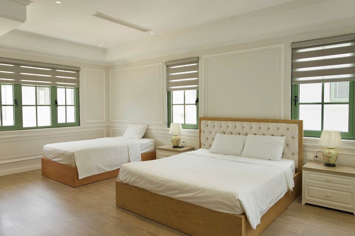 Habitación de hotel con dos camas, un ventanal con persianas, una mesita de noche con lámpara y suelo de madera.