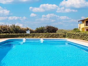 A swimming pool with clear blue water, surrounded by a hedge and a building with yellow walls. - Holiday Home With air Conditioning and Shared Pool (Viddalba)