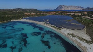 An aerial view of a coastal area with clear blue waters, a sandy beach, and a mountainous backdrop. - Residence Porto Coda Cavallo, Insel Sardinien (San Teodoro)