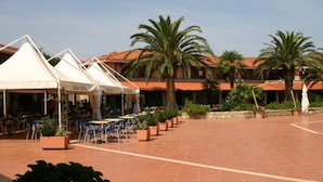 An outdoor patio with tables and chairs, palm trees, and a building with a tiled roof. - Residence Porto Coda Cavallo, Insel Sardinien (San Teodoro)