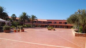 A courtyard with a tiled floor, palm trees, and a building with a red roof. - Residence Porto Coda Cavallo, Insel Sardinien (San Teodoro)
