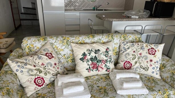A sofa with floral upholstery and a kitchen area in the background. - Residence Porto Coda Cavallo, Insel Sardinien (San Teodoro)