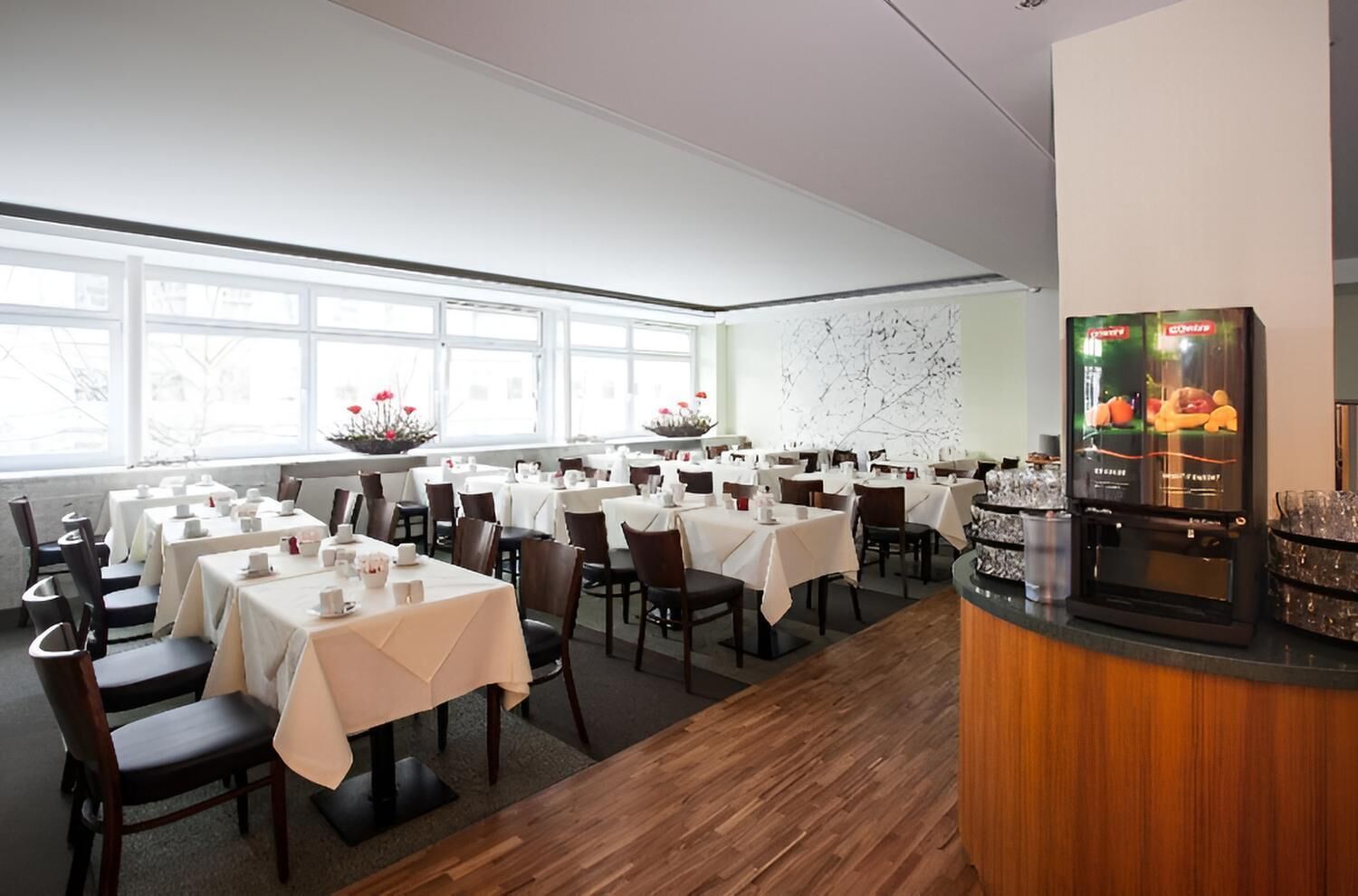 Un restaurant avec des tables dressées pour le service, un coin bar et une grande fenêtre laissant entrer la lumière naturelle.
