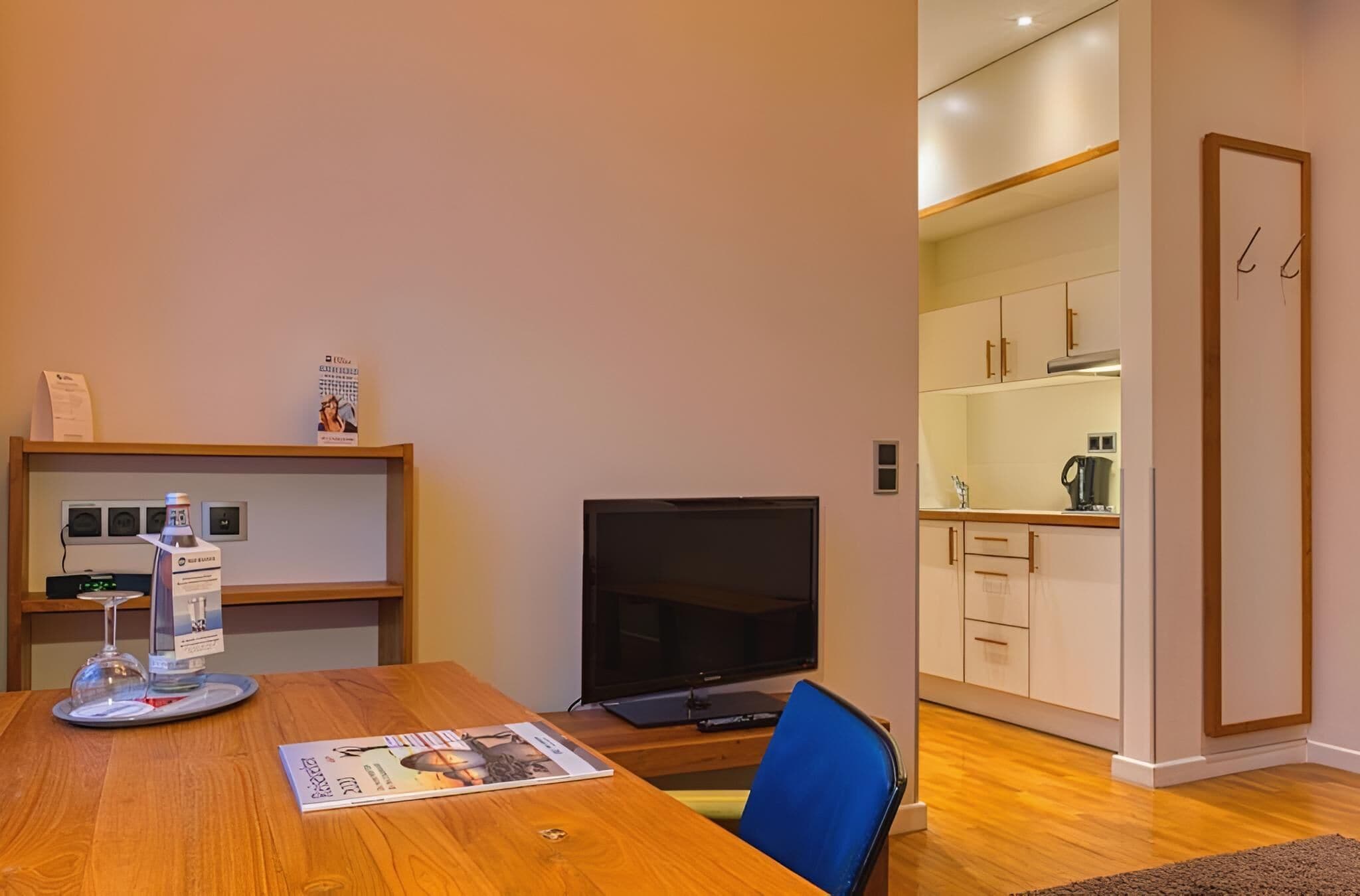 A modern room with a wooden dining table, a flat-screen TV, a magazine, and a shelf with a bottle and a glass.