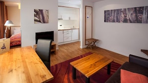 Un salon moderne avec une table à manger en bois, un canapé et un téléviseur. On aperçoit au fond une cuisine avec des meubles blancs et un plan de travail en bois.