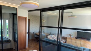 Una habitación de hotel moderna con un amplio ventanal corredero que da a un balcón, una cama con una manta colorida, un televisor sobre un mueble de madera y un área de comedor visible a través de la puerta.