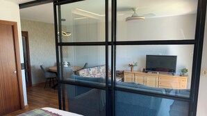 Una habitación moderna con un amplio ventanal corredera, una cama con una manta colorida, un televisor sobre un mueble de madera y un ventilador de techo.