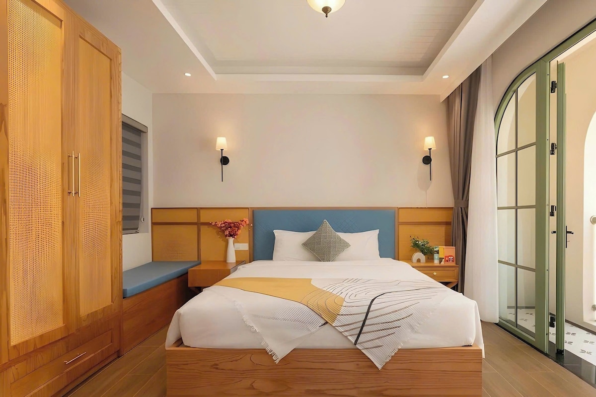 Habitación de hotel con una cama de madera, dos luces fijadas a la pared, una mesita de noche, un florero con flores y vistas a través de una puerta de cristal.