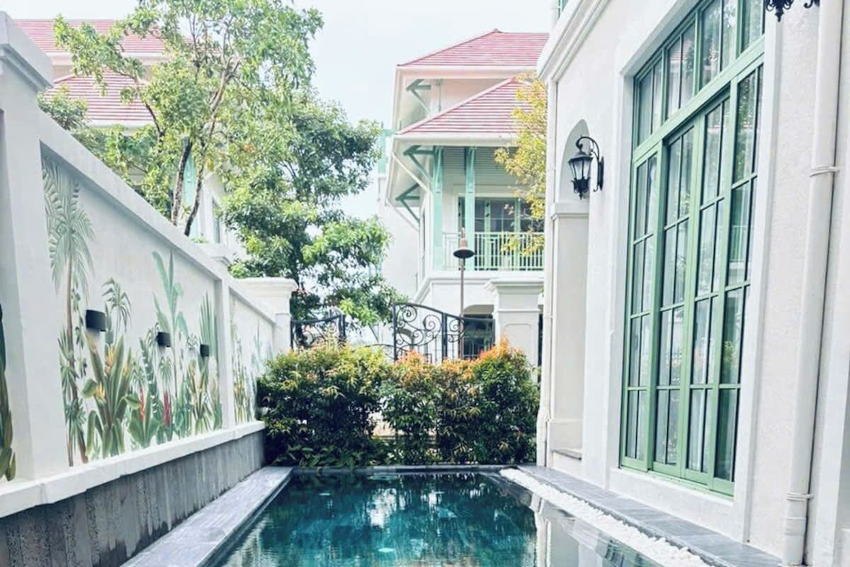 Una piscina con agua cristalina, rodeada por un edificio blanco con marcos de ventanas verdes y un mural de plantas tropicales.