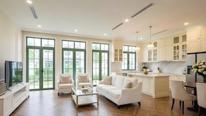 A spacious living room with a sofa, coffee table, and TV, a kitchen with white cabinets, and large windows.