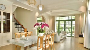 A spacious living area with a dining table, chairs, a staircase, and large windows.
