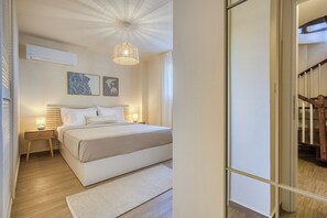 A bedroom with a bed, bedside tables, a hanging light fixture, and a wall-mounted air conditioner. - Bellu - Govino Bay 2BR Maisonette with Common Pool (Gouvia)