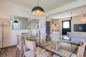 A modern living room with a glass dining table, wooden chairs, a sofa, a TV, and a large mirror. - Bellu - Govino Bay 2BR Maisonette with Common Pool (Gouvia)