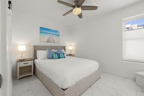 Ein Schlafzimmer mit einem Bett, Deckenventilator, Nachttisch, Nachttischlampe, einem Gemälde und einem Fenster.