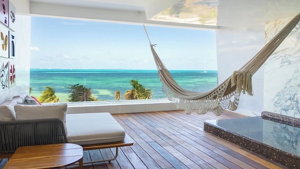 A room with a large window overlooking the ocean, a hammock, a wooden deck, and a seating area with a table. - Luxury Adults-Only Oceanfront Escape in Beautiful Cancun (Cancún)