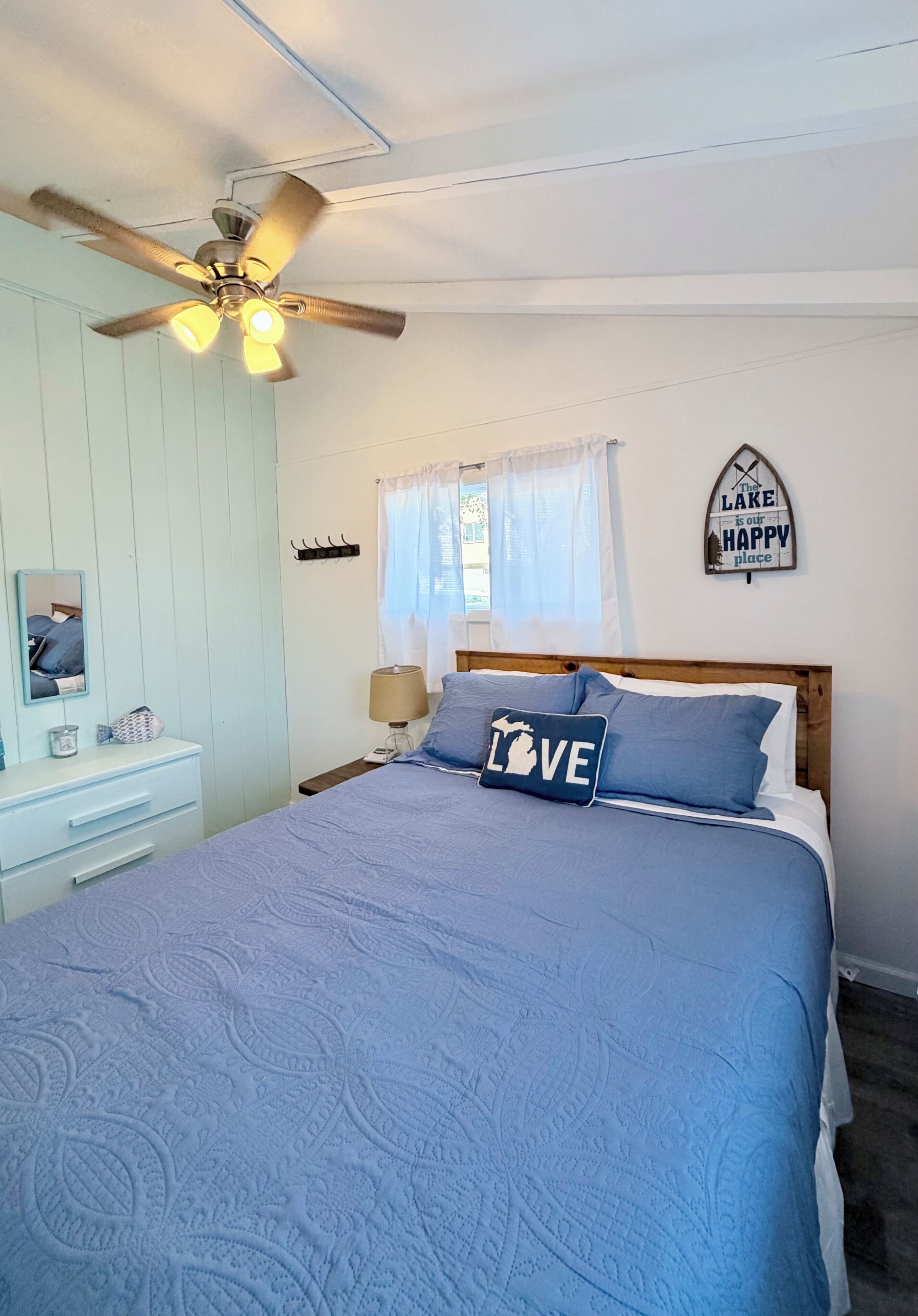 Quarto com cama, ventilador de teto, mesa de cabeceira com abajur, cômoda e um objeto decorativo na parede.