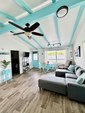 A spacious living area with a ceiling fan, a dining table with chairs, a gray sofa, and a view through the window.