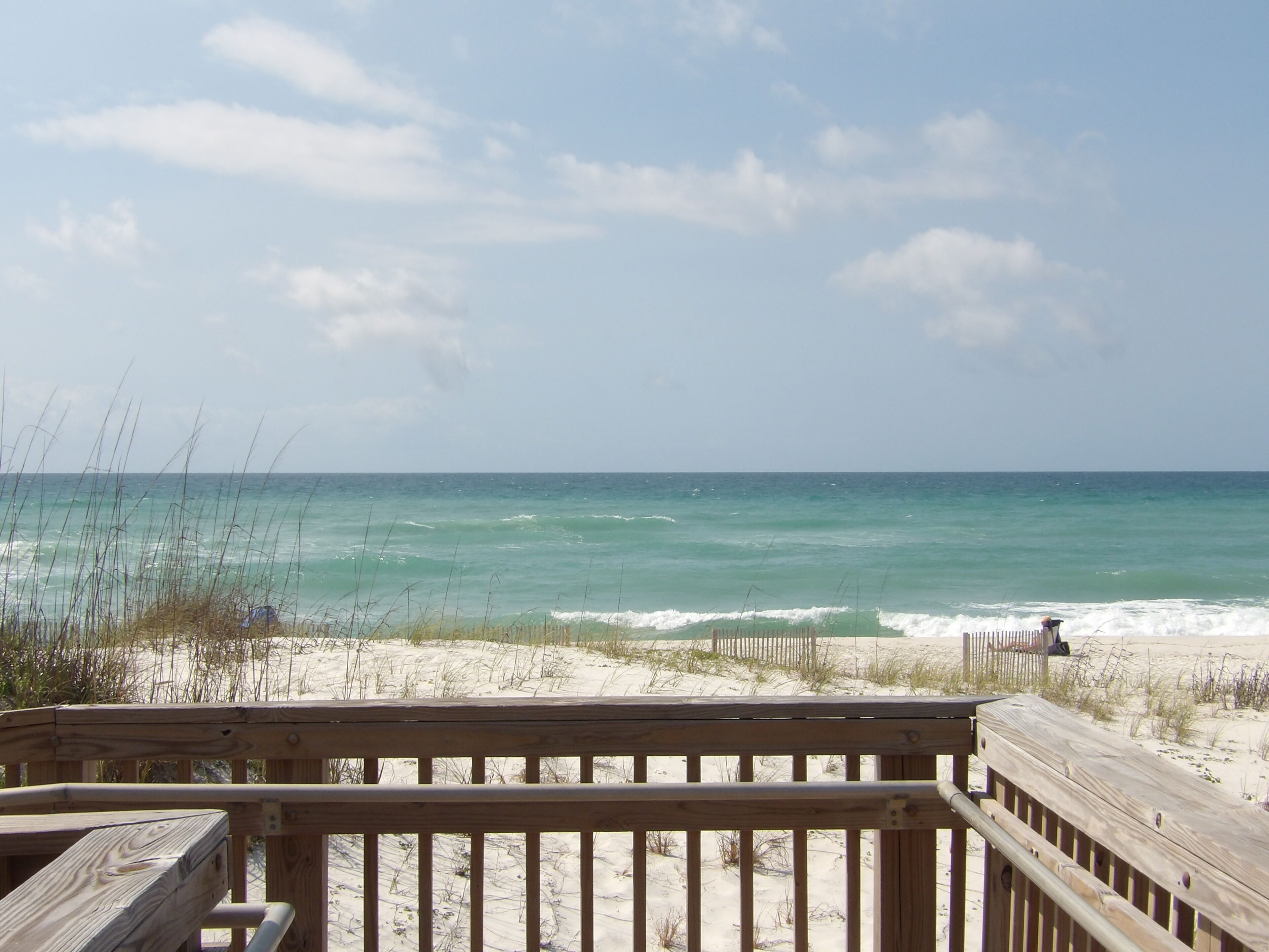 A wooden deck overlooking a beach with waves, sandy dunes, and a clear sky.