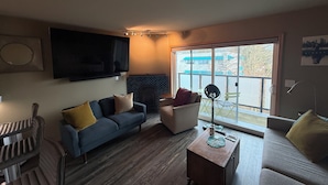 A living room with a sofa, a coffee table, a TV mounted on the wall, and a view through a large window. - Modern 1-bedroom condo in West Seattle, with quick access to downtown (Seattle)