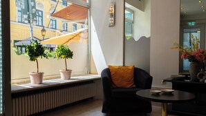 A modern interior with a black sofa, a small round table, potted plants, and a view of the street outside. - Hotel Mondial Munich (Munich)