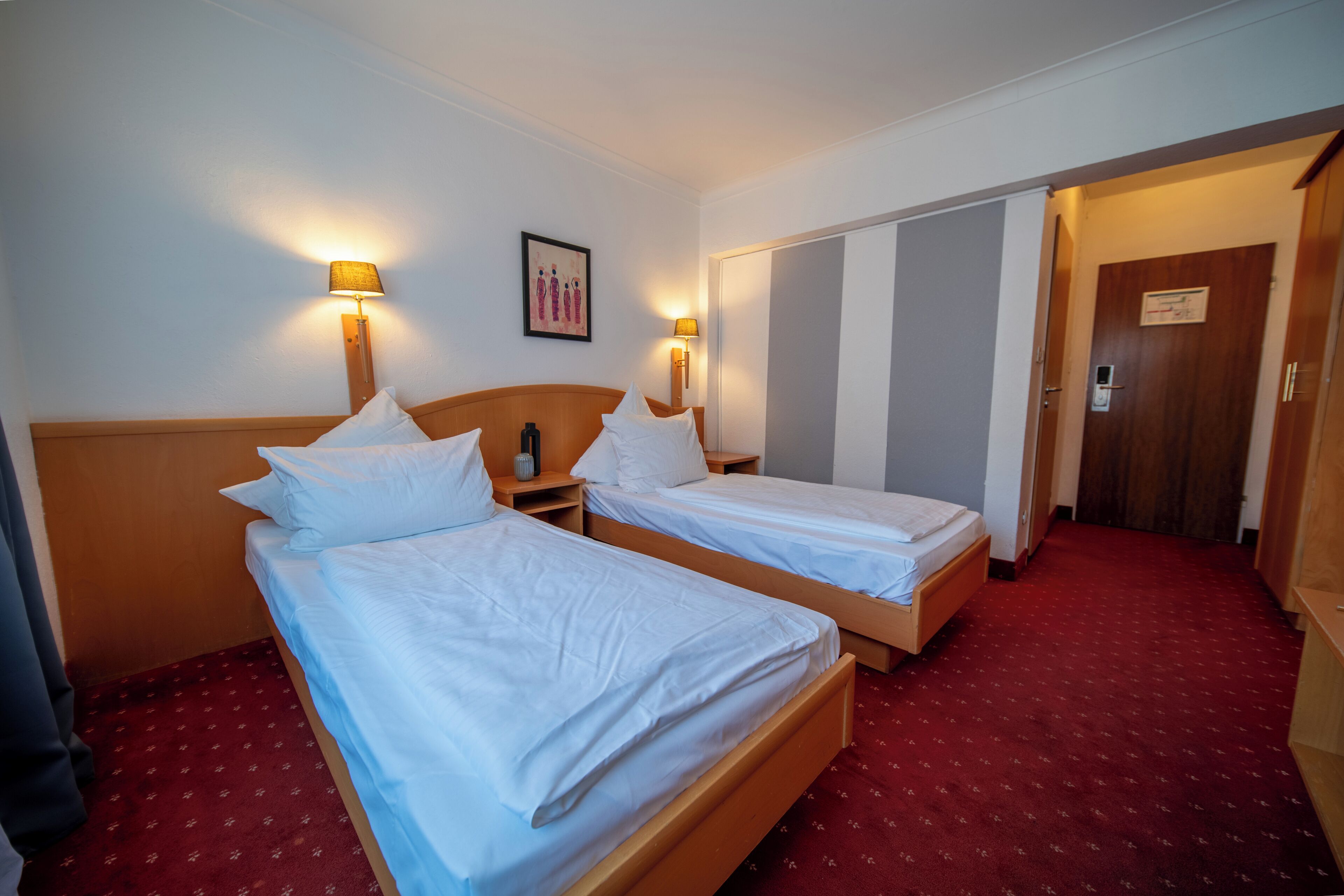 A hotel room with two single beds, a wooden headboard, bedside lamps, a red carpeted floor, and a door leading to another room.