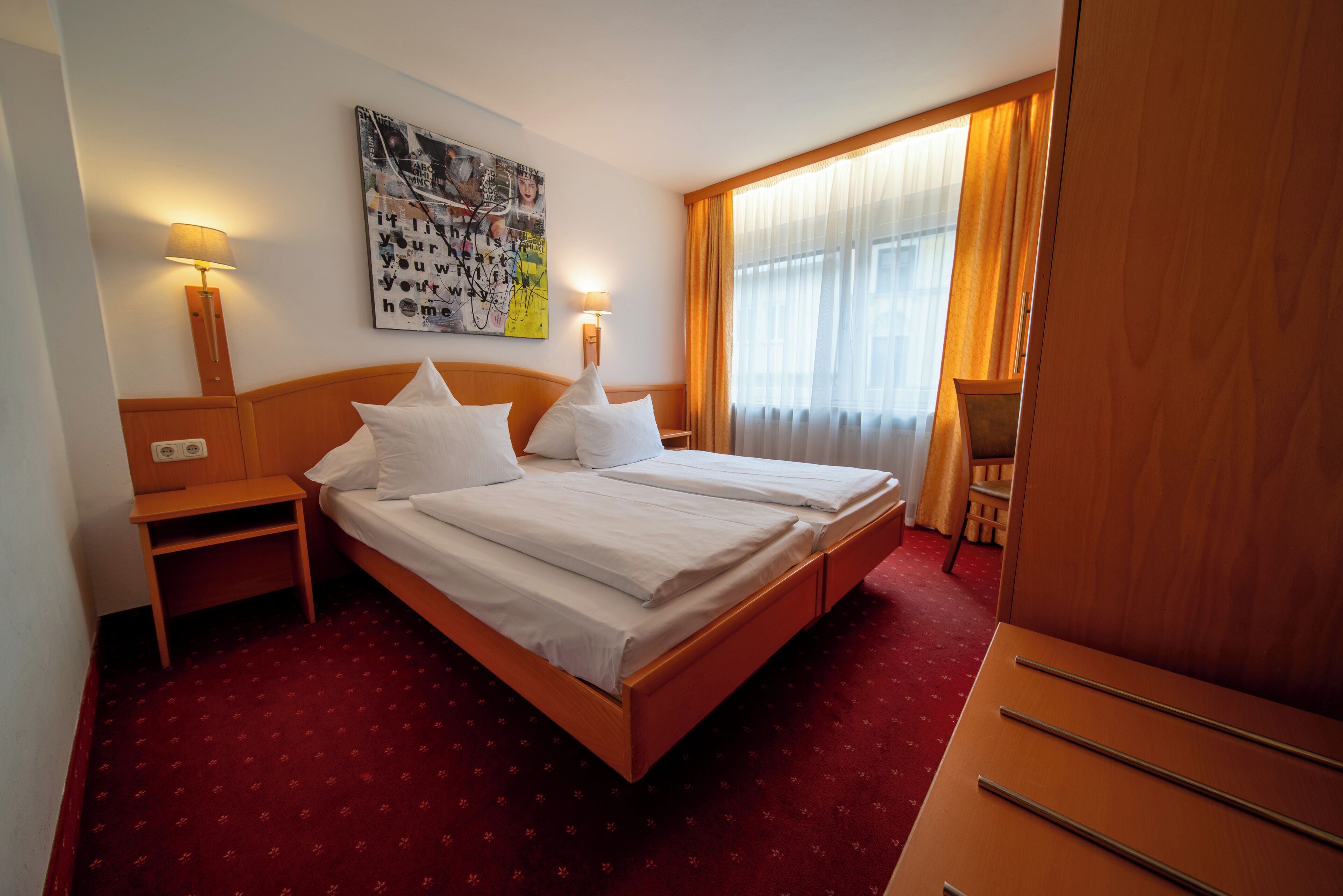 Une chambre d’hôtel avec un lit double, deux tables de chevet, une petite table, une lampe fixée au mur, une fenêtre avec des rideaux et une armoire en bois.