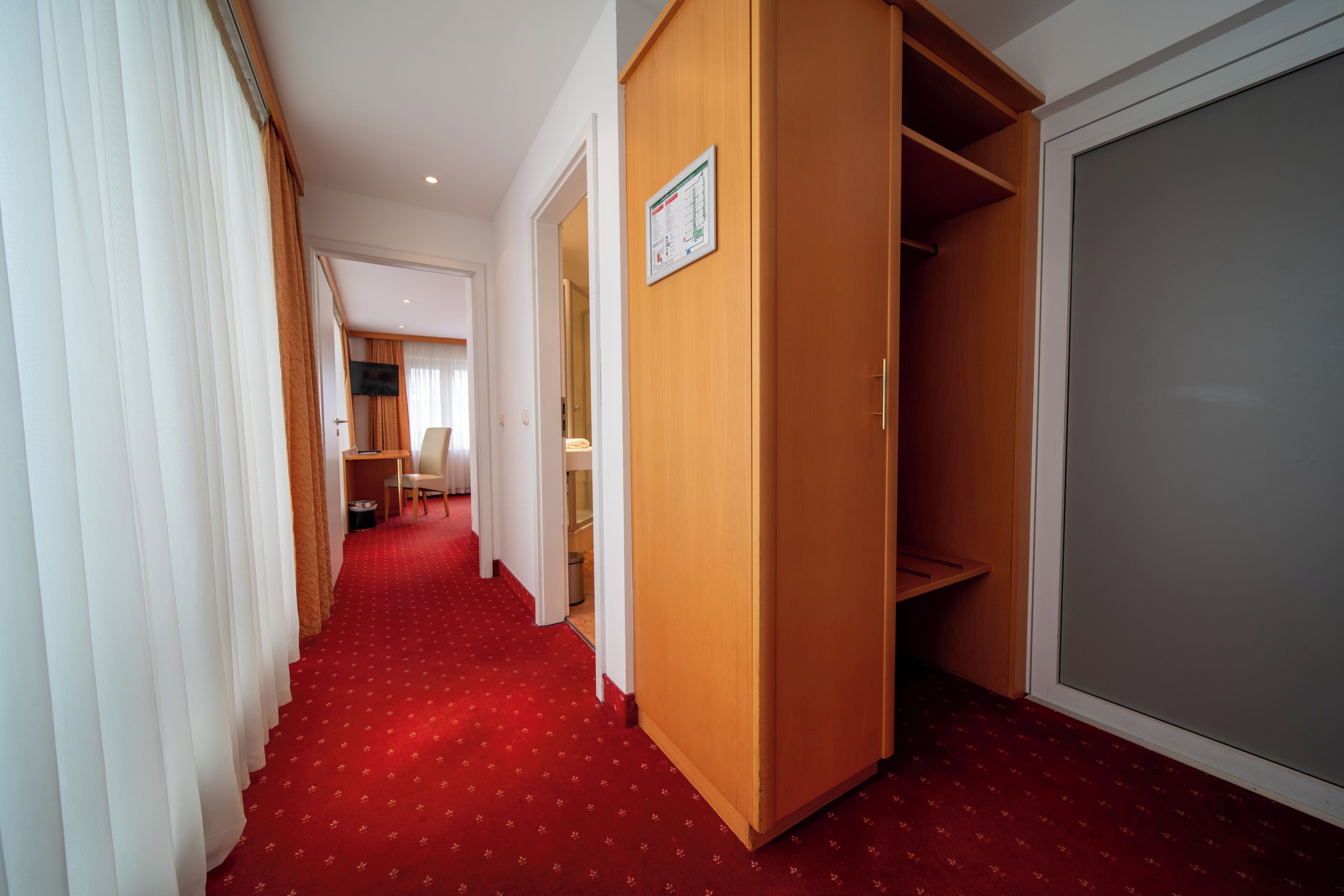 A hotel corridor with a red carpet, wooden wardrobe, and a closed door.