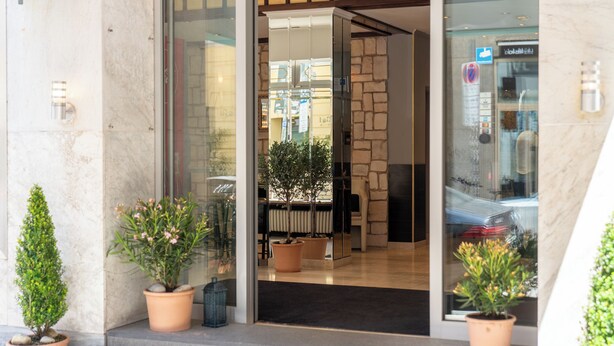 A hotel entrance with a stone wall, potted plants, and a glass door. - Hotel Mondial Munich (Munich)