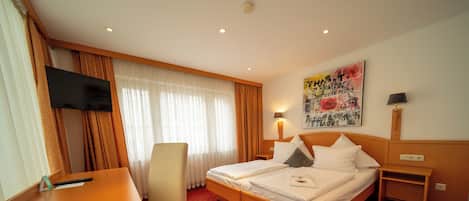 A hotel room with a bed, a desk, a chair, a TV, a painting, and a lamp.