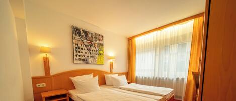 A hotel room with a double bed, a nightstand, a lamp, a painting on the wall, and a window with curtains.