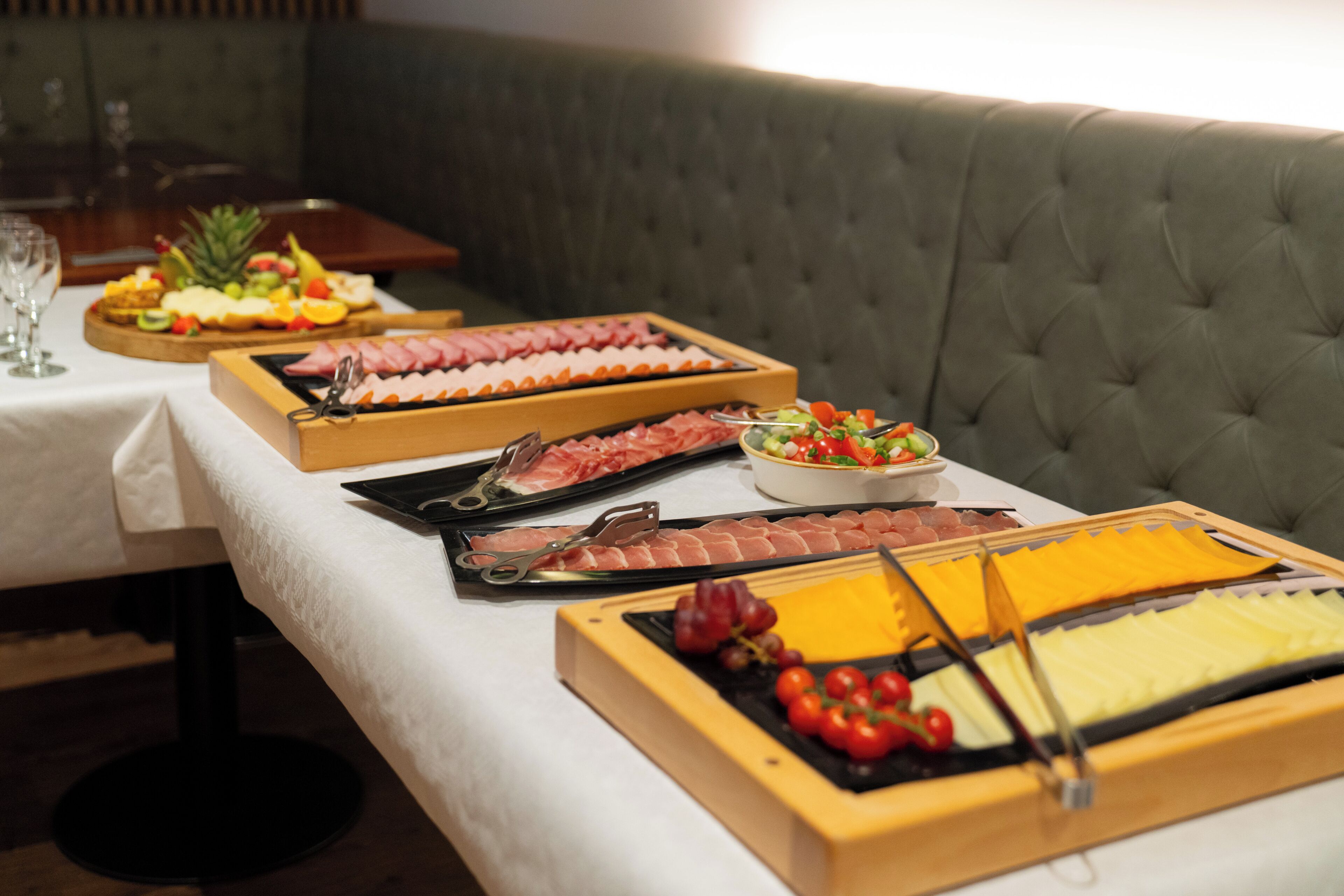 Una mesa de buffet con diversas opciones de comida, entre ellas frutas, quesos y embutidos, sobre un mantel blanco en un comedor con un sofá verde capitonado al fondo.