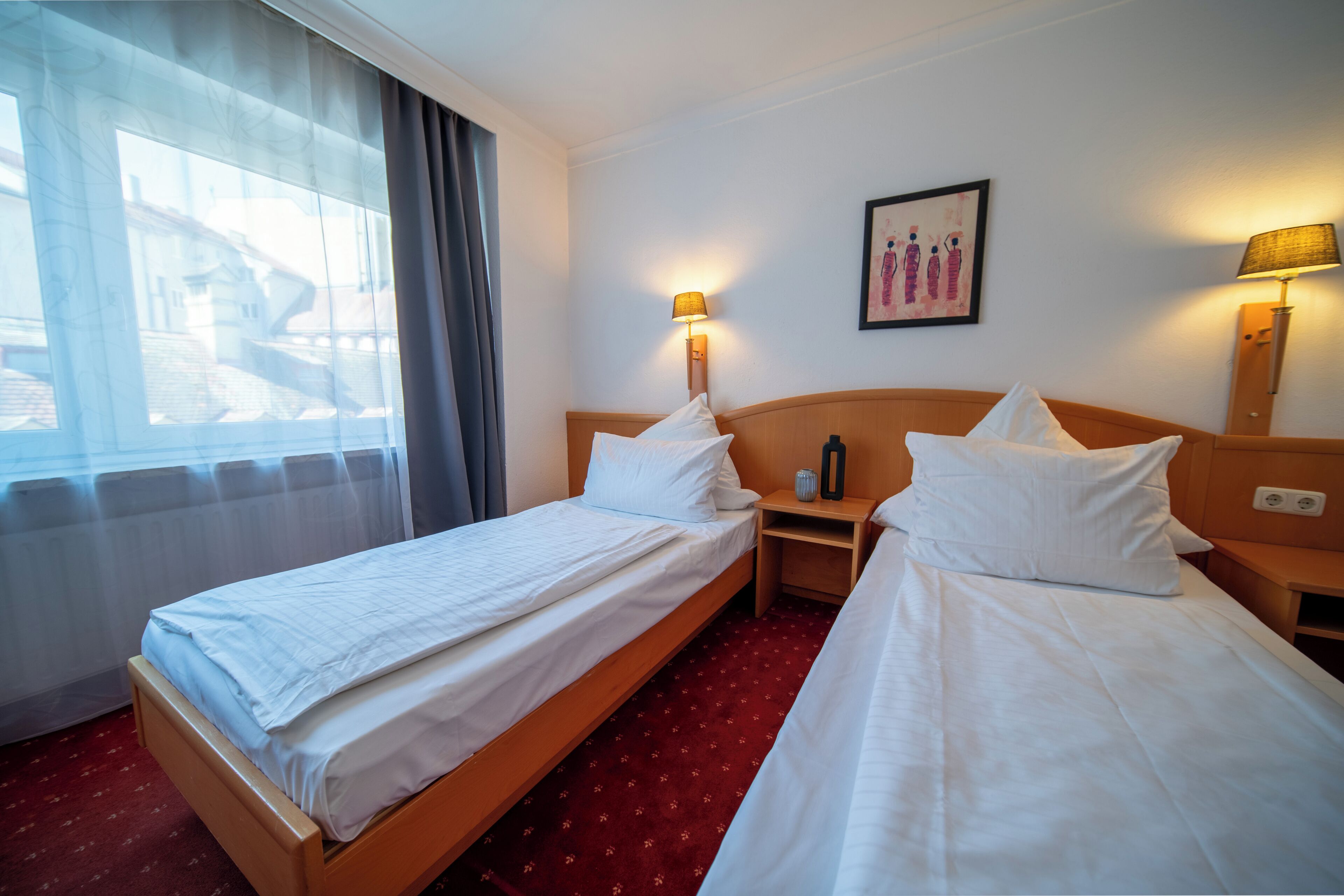 A hotel room with two single beds, a window with curtains, a wall-mounted lamp, and a framed picture.