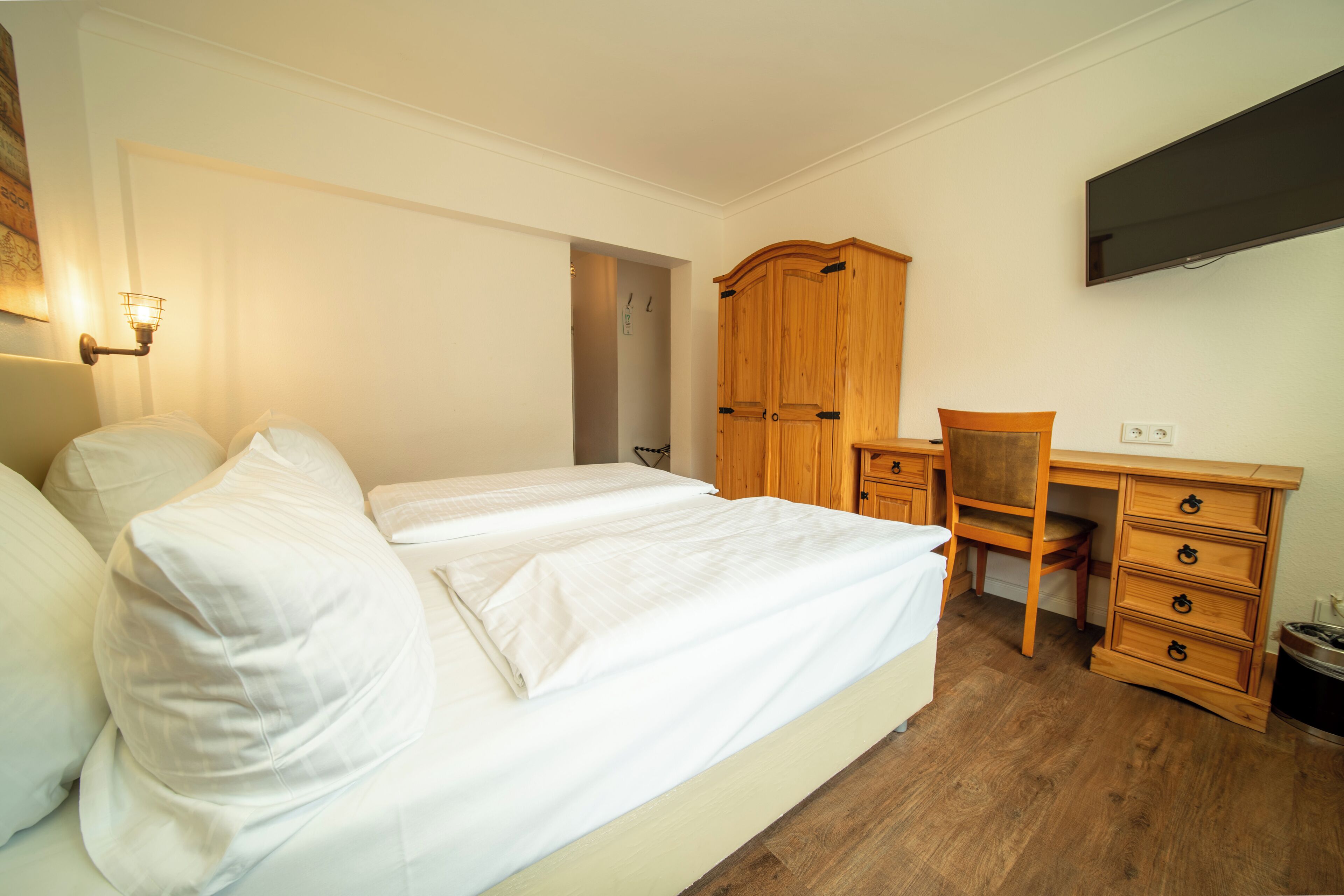A bedroom with a bed, a wooden desk, a chair, a wardrobe, and a flat-screen TV mounted on the wall.