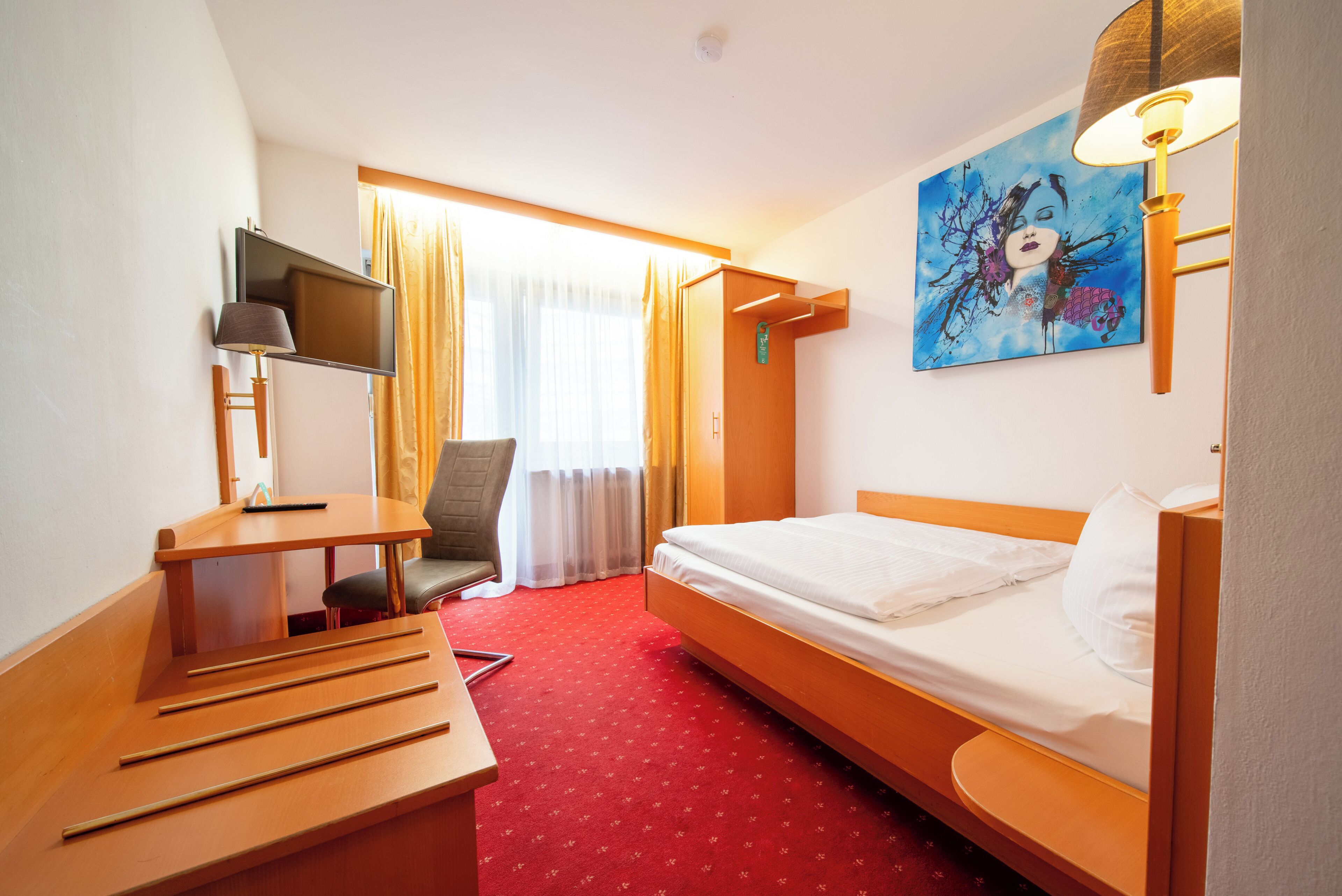 A hotel room with a bed, desk, chair, TV, and artwork on the wall.