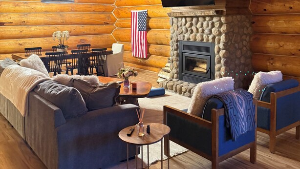 A log cabin interior with a fireplace, a television mounted on the wall, a dining area, and a cozy seating arrangement.