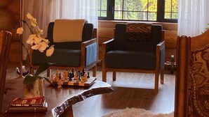 A log cabin interior with two chairs, a chessboard, a lamp, and a view of autumn trees through large windows.
