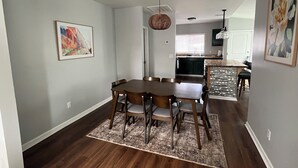A dining area with a wooden table and chairs, a kitchen in the background, and a hanging light fixture.