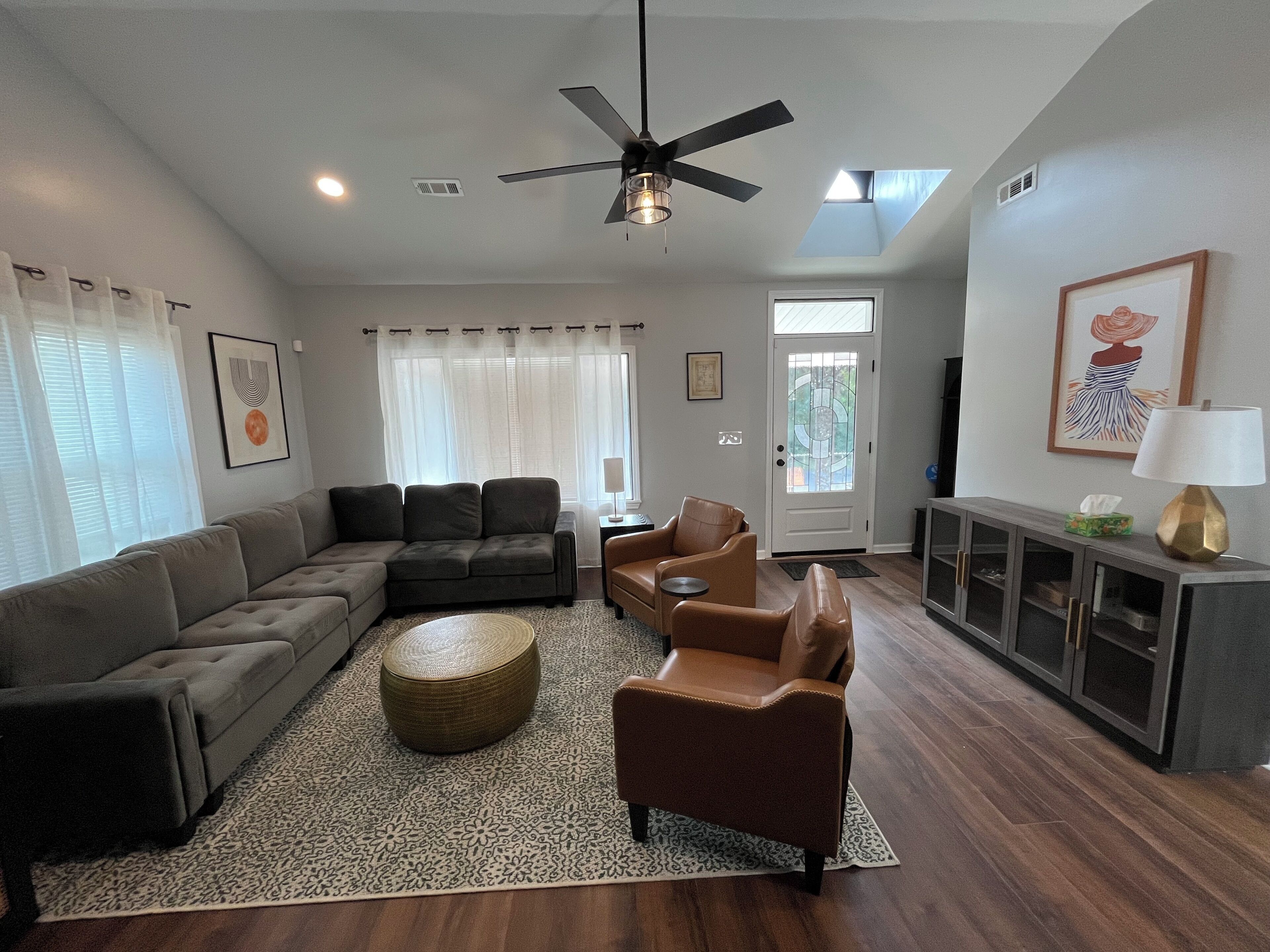 A living room with a sectional sofa, a brown armchair, a round ottoman, a ceiling fan, and a TV on a cabinet.