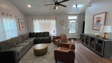 A living room with a sectional sofa, a brown armchair, a round ottoman, a ceiling fan, and a TV on a cabinet.